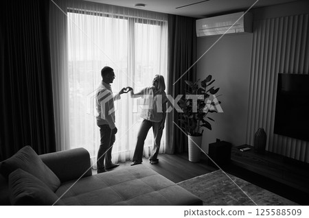 Lovely couple showing heart symbol. Young man and his girlfriend standing opposite the window in natural light at home. Cute newlyweds happy about their new apartment. Black and white image. Lovely couple showing heart symbol. Young man and his girlfriend standing opposite the window in natural light at home. Cute newlyweds happy about their new apartment. Black and white image. 125588509