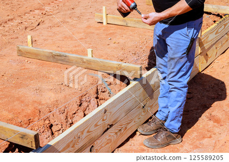 Construction worker uses drill to secure wooden forms on work day at construction site for foundation 125589205