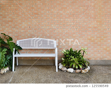 A clean white metal bench is placed in front of the brick-walled. 125590549