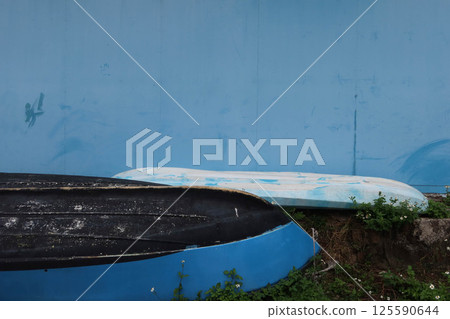 two boats old weathered positioned at the angle resting against a blue wall, depicts a serene backdrop, and patches of greenery and small flowers around the boats with copy space two boats old weathered positioned at the angle resting against a blue wall, depicts a serene backdrop, and patches of greenery and small flowers around the boats with copy space 125590644