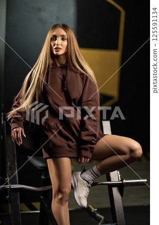 young woman long hair stands in gym setting dressed in cozy brown hoodie and athletic shorts. Her confident pose shows dedication fitness and overall wellness. young woman long hair stands in gym setting dressed in cozy brown hoodie and athletic shorts. Her confident pose shows dedication fitness and overall wellness. 125591134