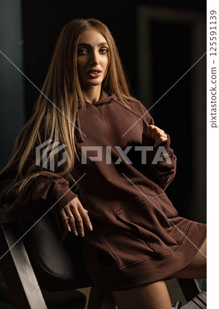woman long hair poses confidently while seated on chair dressed in brown hoodie. studio's soft lighting highlights her features and creates intriguing atmosphere. woman long hair poses confidently while seated on chair dressed in brown hoodie. studio's soft lighting highlights her features and creates intriguing atmosphere. 125591139