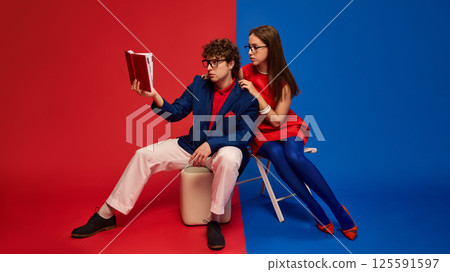 Stylish man in glasses, sitting and reading book to woman sitting close behind against split red and blue background. 125591597