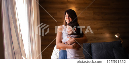 Portrait of Caucasian mom and daughter hug and cuddle in living room standing by window. Understanding relationships, love and family bonding concept. 125591832