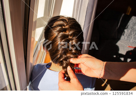 Close-up of mother's hands weaving braid in daughter's hair. Beauty girly routine before school and feminine bonding. Authentic parenting moment. 125591834