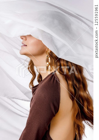 woman with long wavy red hair and golden earrings smiling under white sheer fabric in brown open-back dress, feminine mood in studio scene, fashion, jewelry design, beauty editorial 125591961