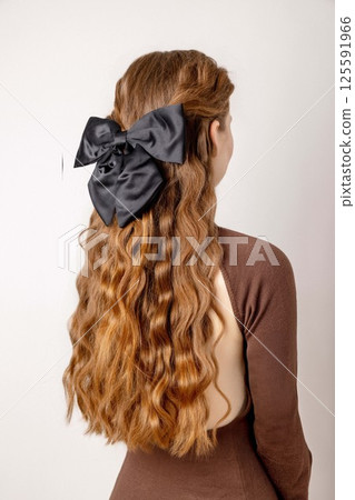 young woman with long wavy red hair styled with large black bow accessory, side rear view in minimalist bright studio with neutral background, hair fashion, beauty salon, hair accessories 125591966