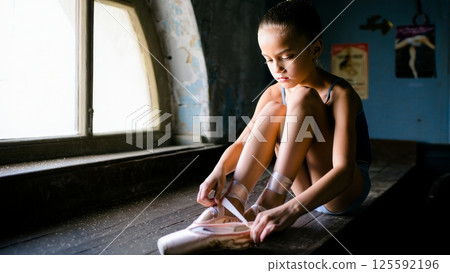 A young ballerina ties the ribbons of her pointe shoes while sitting on a weathered wooden bench beside a large arched window A young ballerina ties the ribbons of her pointe shoes while sitting on a weathered wooden bench beside a large arched window 125592196