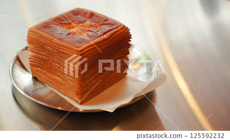A cube-shaped mille crepe cake rests on a silver plate, each golden-brown crepe layer stacked with precise uniformity. The soft lighting reflects off the metal surface of the table, highlighting the 125592232