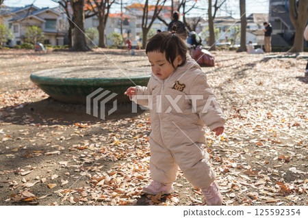 [One-year-old child standing and playing in a park with fallen winter leaves] 125592354