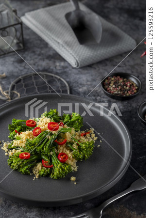 Gourmet dish of tender stem broccoli, couscous and chili pepper on black plate on a dark background Gourmet dish of tender stem broccoli, couscous and chili pepper on black plate on a dark background 125593216