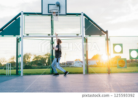 Young Man with basketball ball under hoop, Playing Basketball at Park outdoor playground in sunset light with blue sky background. Basketball match. Teenagers' hobby. Sports and healthy lifestyle 125593392