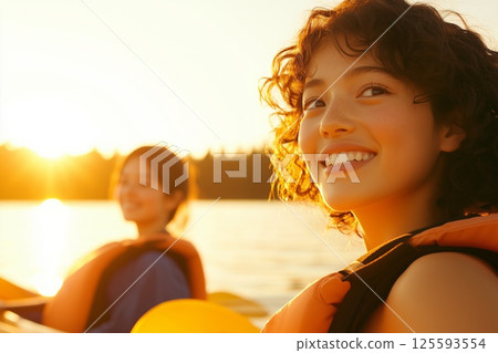 Two women enjoying a peaceful kayaking break in the afternoon sun by the calm water and forest 125593554