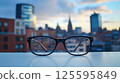 Pair of glasses is on a table in front of a city skyline. The glasses are black and have a reflective surface. The cityscape in the background is a mix of tall buildings and clouds 125595849
