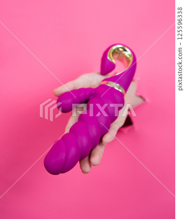 Female hand sticking out with pink sex toy from pink paper background. Vertical photo. Female hand sticking out with pink sex toy from pink paper background. Vertical photo. 125596338
