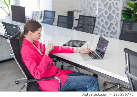 Caucasian business woman in fuchsia jacket drinking coffee at her desk.  125596773