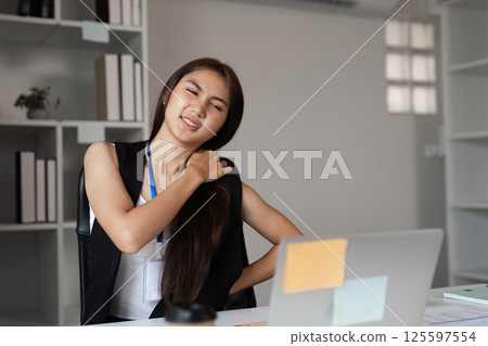 Young Businesswoman Experiencing Muscle Aches at Work in Modern Office Setting with Laptop and Documents Young Businesswoman Experiencing Muscle Aches at Work in Modern Office Setting with Laptop and Documents 125597554