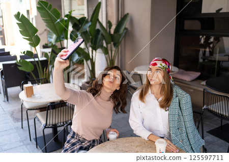 Two young women sitting at an outdoor cafe , enjoying coffee and taking a selfie together. Casual moment of friendship and lifestyle. 125597711
