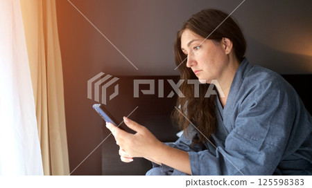 Female tourist sits on bed edge focused on phone in hotel apartment Female tourist sits on bed edge focused on phone in hotel apartment 125598383