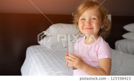 Little girl drinks water from clear glass perched on bed edge Little girl drinks water from clear glass perched on bed edge 125598423