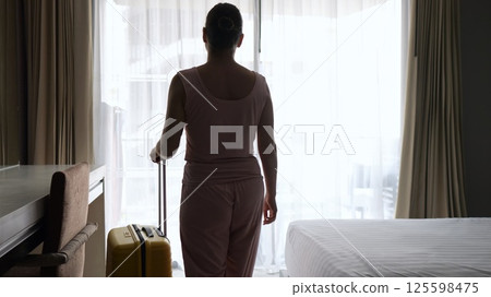 Woman in pink pyjamas walks toward window in morning 125598475