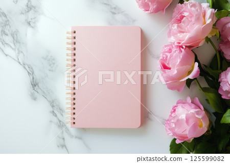 Soft pink notebook with spiral binding placed on elegant marble surface. Beautiful blooming peonies on the side create romantic and gentle flat lay. Ideal for feminine design or planning themes. 125599082