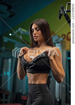 fitness enthusiast stands in gym showing her dedication strength training. She adjusts her top while showcasing her toned physique and confidence. 125599695