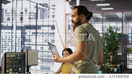 Cheerful admin holding laptop chatting with colleagues while working with machine learning tech. Man transforming data into predictive models using notebook and talking with coworkers, camera A 125599708