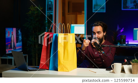 Excited media star holding colorful shopping bags, showing fans purchases, filming himself in apartment. Playful content creator in studio presenting audience new acquisitions, camera A Excited media star holding colorful shopping bags, showing fans purchases, filming himself in apartment. Playful content creator in studio presenting audience new acquisitions, camera A 125599714