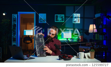 Man in studio reviewing gaming keyboard received from sponsoring brand. Online show host paid to promote bluetooth wireless computer peripheral to viewers during live broadcast, camera B Man in studio reviewing gaming keyboard received from sponsoring brand. Online show host paid to promote bluetooth wireless computer peripheral to viewers during live broadcast, camera B 125599727