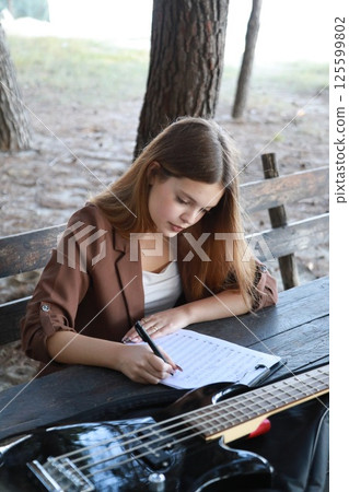 Girl Writes Music Notes On Sheet Music Sitting At The Park Table In The Nature 125599802