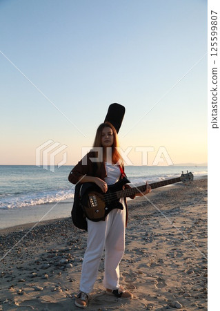 Girl Walks And Plays The Bass Instrument On The Beach Near The Ocean At Sunset 125599807