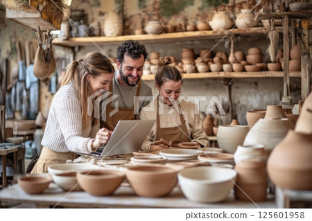 Group of artisans engaged in pottery making while using a laptop for guidance in a workshop setting 125601958
