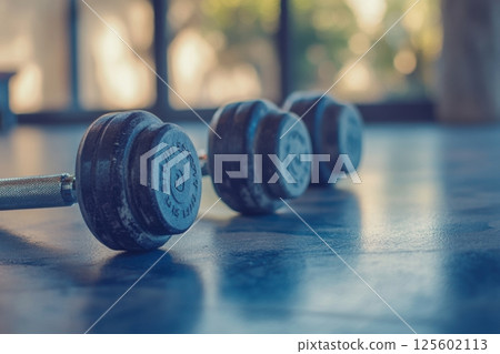 Dumbbells rest on a gym floor in a sunlit fitness center during early morning hours 125602113