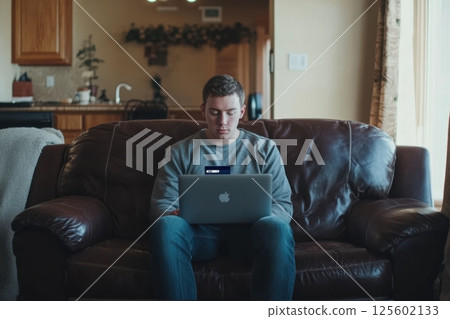 Young man sitting on couch working on laptop in cozy living room during daytime 125602133