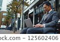 A businessman works on his laptop while sitting on a bench outdoors. 125602406