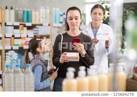Woman with daughter in pharmacy scans QR code on tablet box, pharmacist in background Woman with daughter in pharmacy scans QR code on tablet box, pharmacist in background 125604279