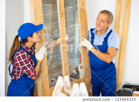 Two contractor repairmen in a blue uniform holding a door in polyethylene before installation in the house 125604333