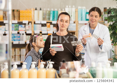 Woman with schoolgirl daughter are busy choosing tablet box at pharmacy, pharmacist in background Woman with schoolgirl daughter are busy choosing tablet box at pharmacy, pharmacist in background 125604341