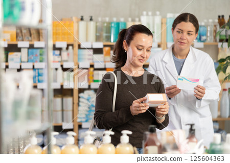 Woman with schoolgirl daughter are busy choosing tablet box at pharmacy, pharmacist in background 125604363