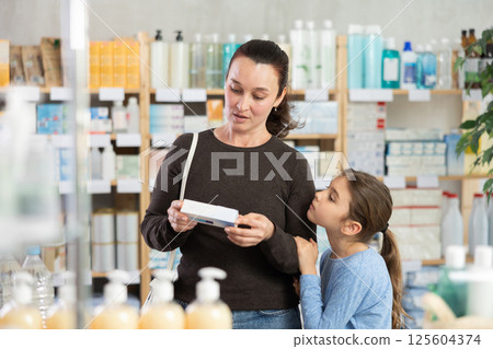 Woman with schoolgirl daughter are busy choosing ointment at pharmacy 125604374