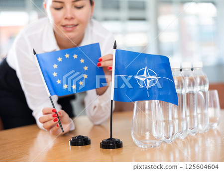 Female secretary places flags of the NATO (OTAN) and European Union flag on the table before negotiations of top political figures Female secretary places flags of the NATO (OTAN) and European Union flag on the table before negotiations of top political figures 125604604