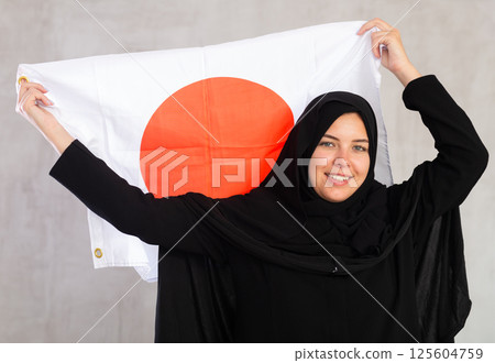 Smiling joyful Muslim woman in traditional black hijab holds flag of Japan 125604759