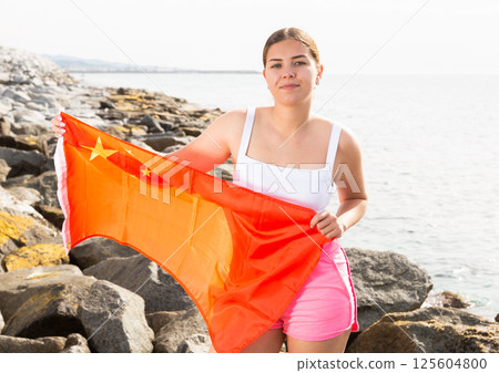 Young woman holding China waving flag at coastline against of bright sea 125604800