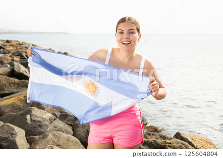 Portrait of happy woman of Argentina match waving a national flag on the seashore on sunny day 125604804