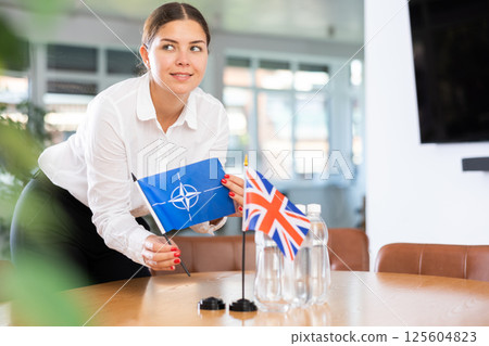 Positive young woman putting little flag of NATO on table next to the flag of Great Britain 125604823