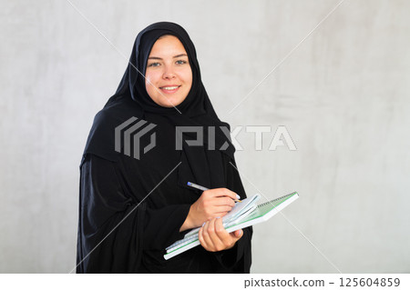 Cheerful young muslim student in black traditional hijab with notebooks in hands 125604859