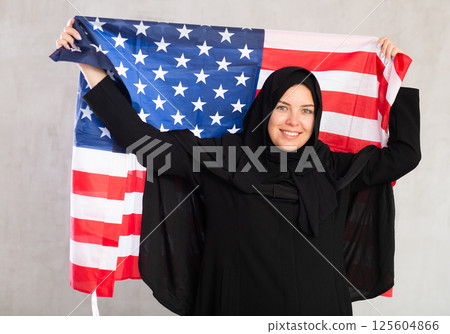 Balanced smiling Muslim woman in traditional black hijab holds flag of United States of America 125604866