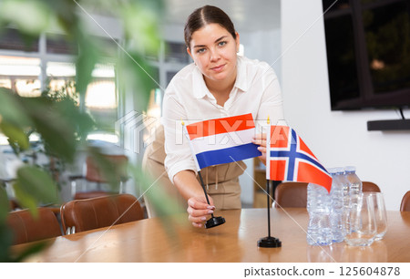 Positive young woman putting flag of Holland on the table with flag of Norway in conference room Positive young woman putting flag of Holland on the table with flag of Norway in conference room 125604878