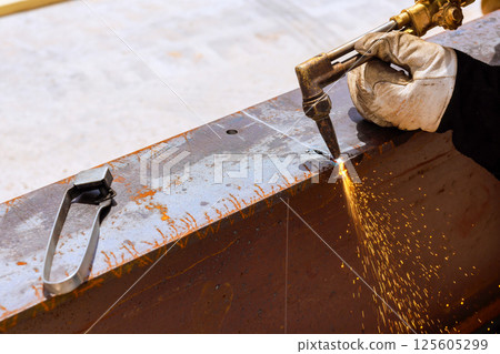 Worker skillfully cuts through metal beam using welding torch, generating sparks at construction site. Worker skillfully cuts through metal beam using welding torch, generating sparks at construction site. 125605299